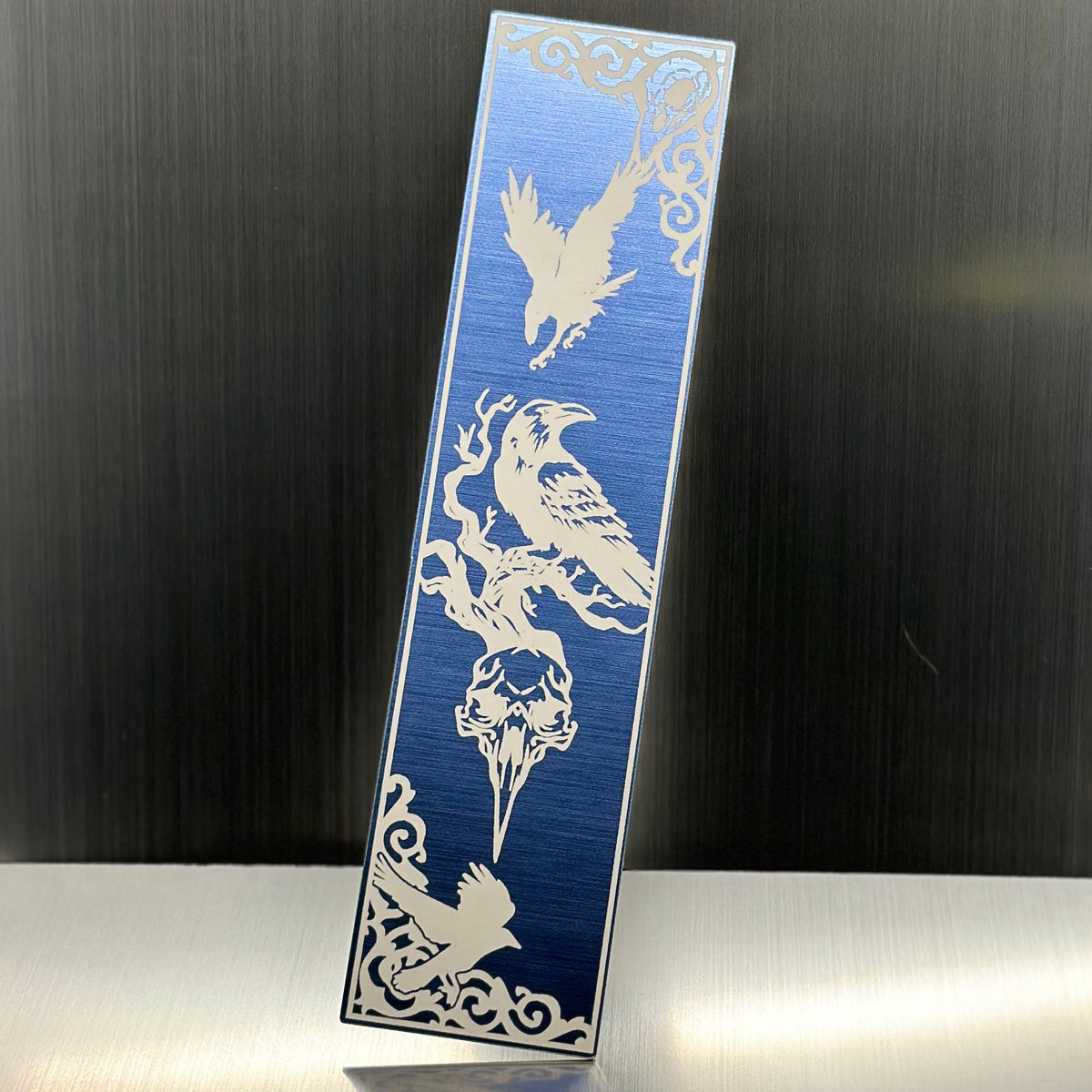 A blue metallic acrylic bookmark with crows and a bird skull.