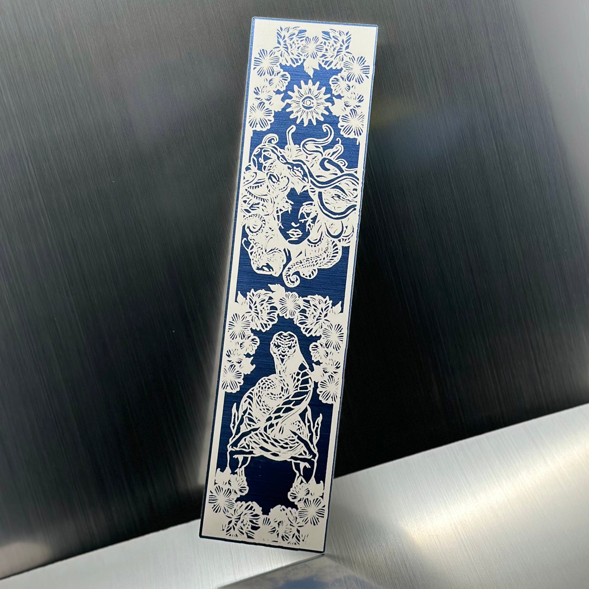A blue metallic acrylic bookmark with Medusa's head at the top and a snake being held at the bottom