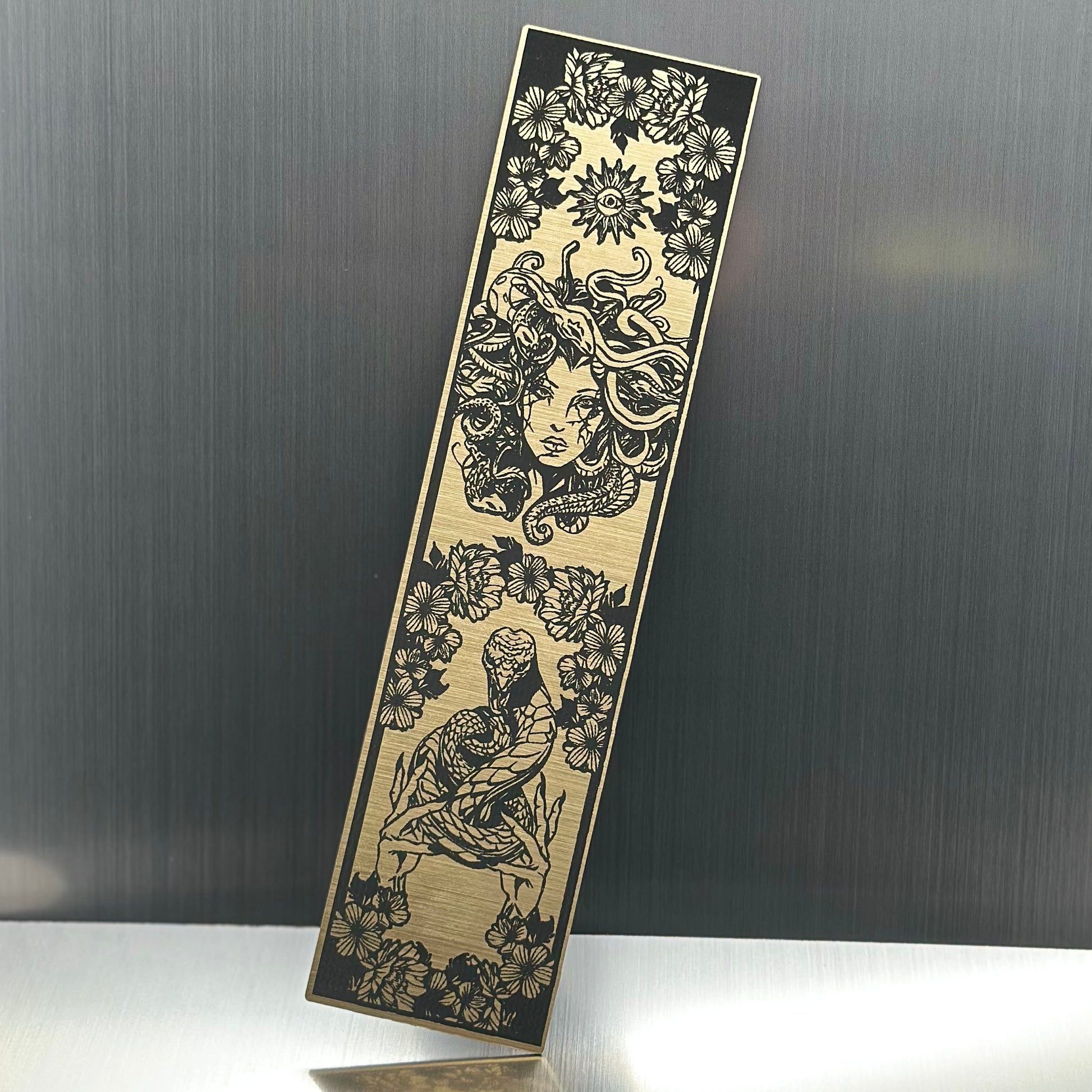 A gold metallic acrylic bookmark with Medusa's head at the top and a snake being held at the bottom
