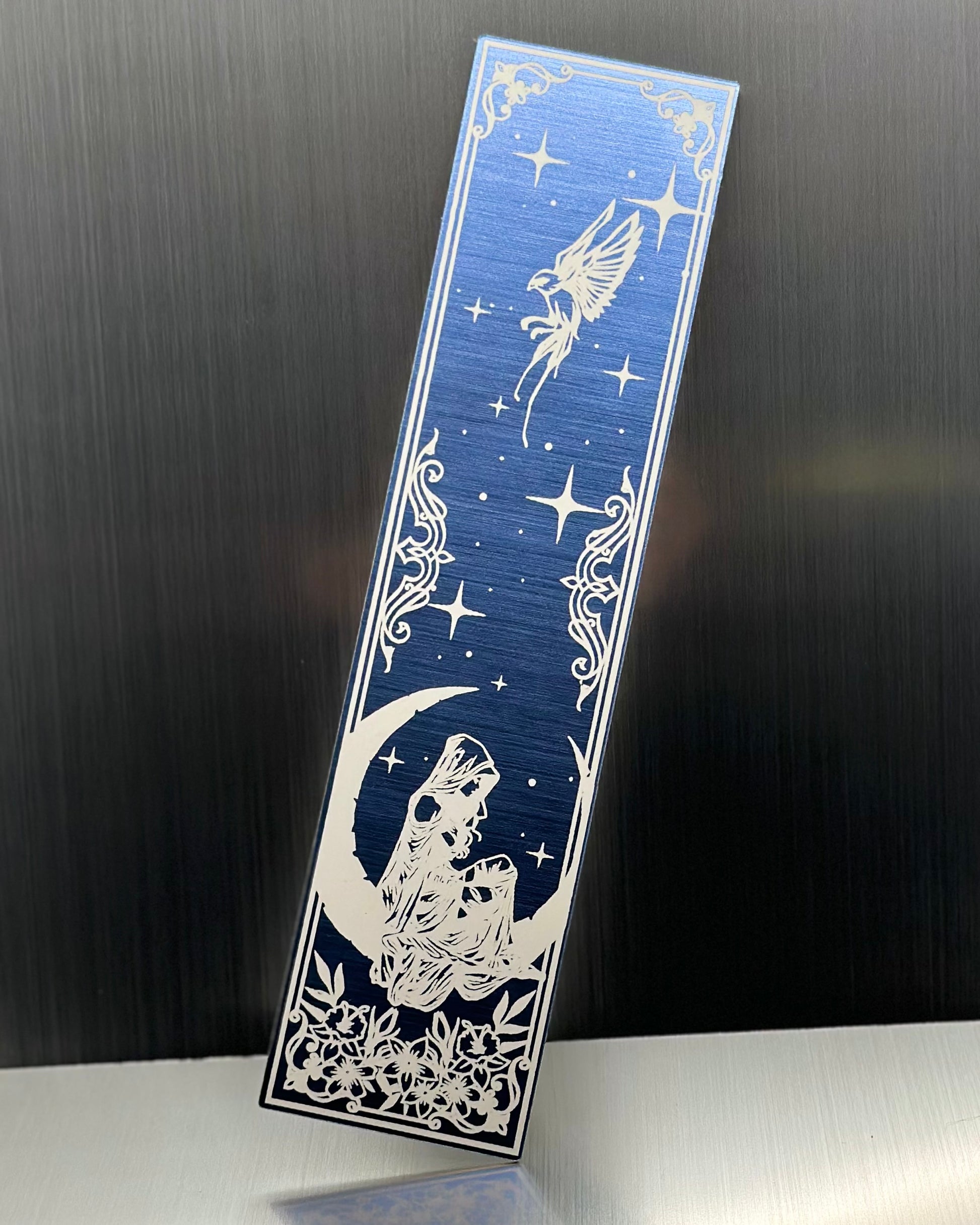 A blue metallic acrylic bookmark with a heart with a feminine person sitting on a crescent moon with a sparrow at the top