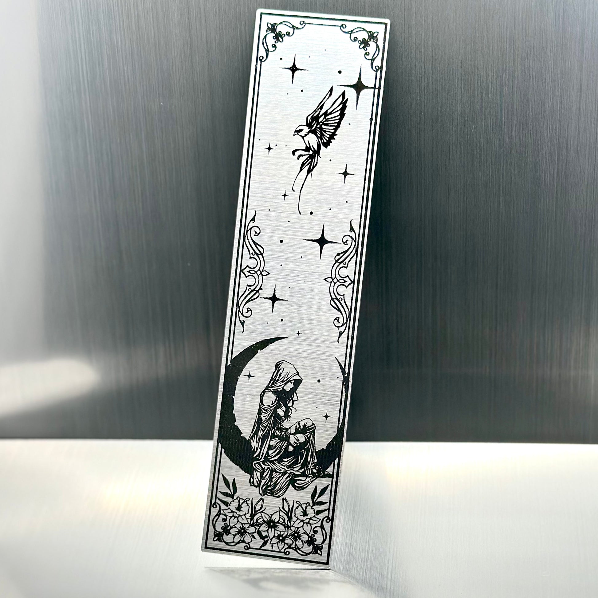 A silver metallic acrylic bookmark with a heart with a feminine person sitting on a crescent moon with a sparrow at the top