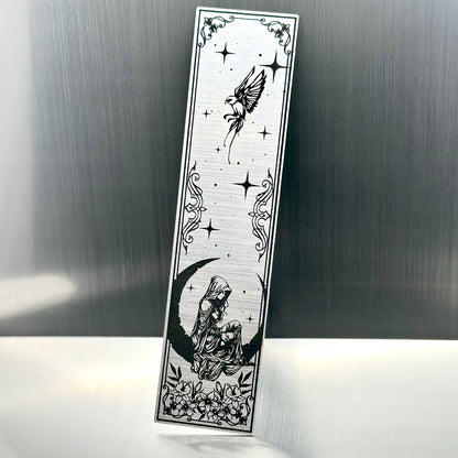 A silver metallic acrylic bookmark with a heart with a feminine person sitting on a crescent moon with a sparrow at the top