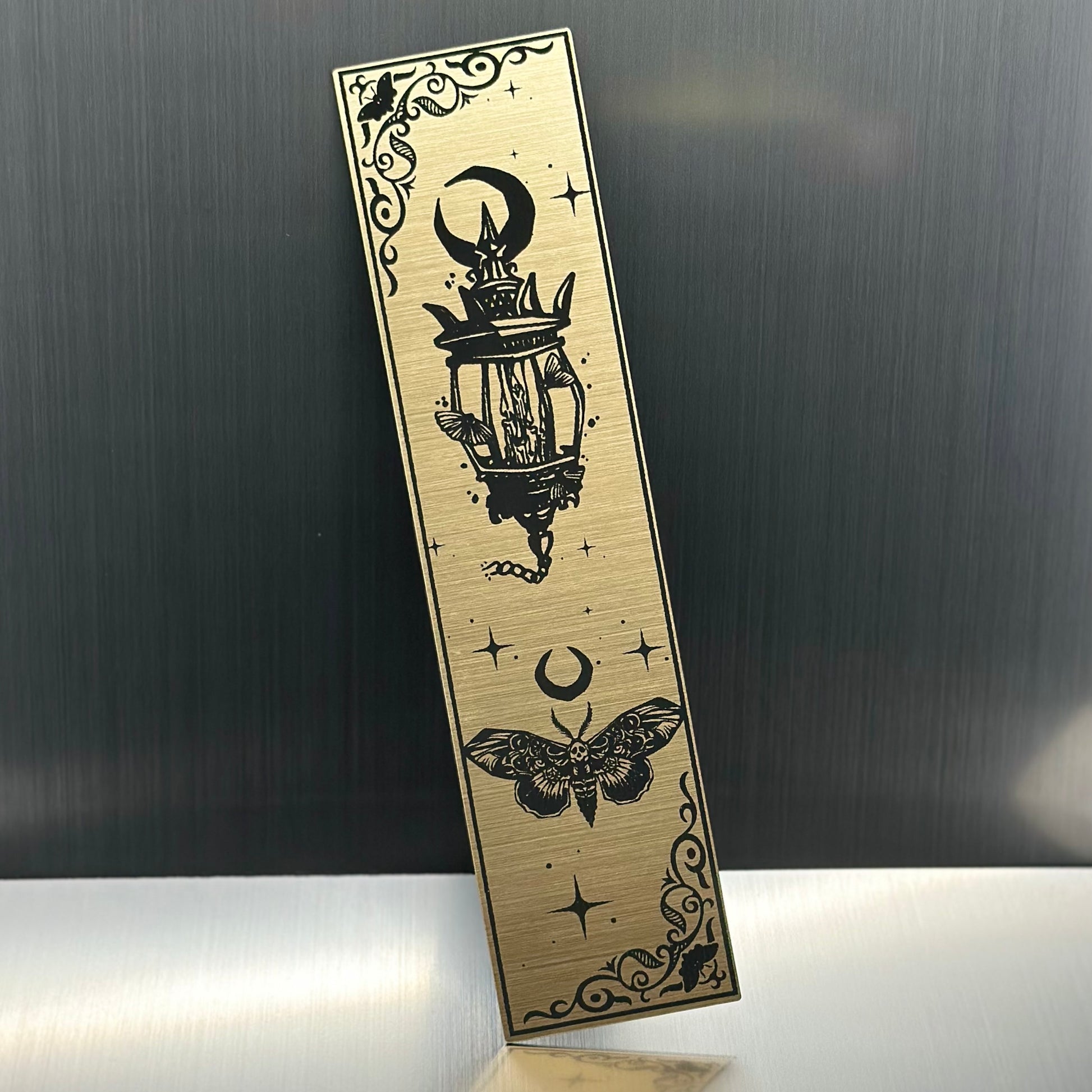 A gold metallic acrylic bookmark with a lantern with a trio of moths