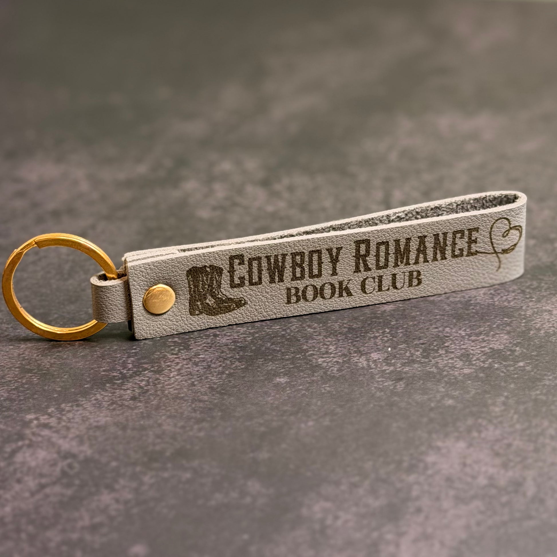 An eggshell white faux-leather keychain with 'Cowboy romance book club' etched into it.