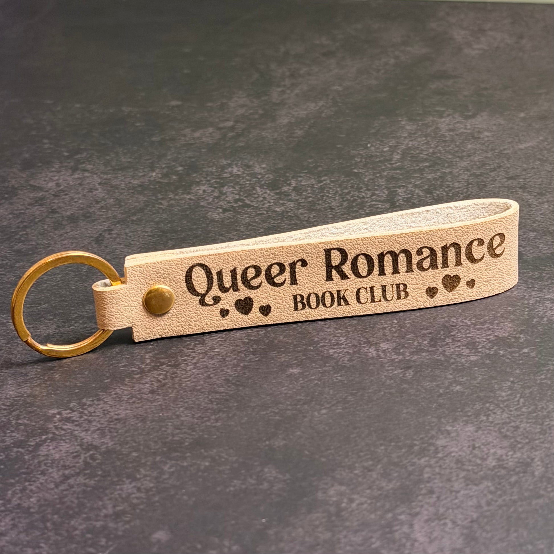 A beige blue faux-leather keychain with 'queer romance book club' etched into it.