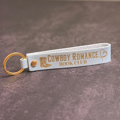 A sky blue faux-leather keychain with 'Cowboy romance book club' etched into it.