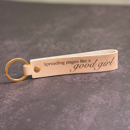 A beige faux-leather keychain with 'Spreading pages like a good girl' etched into it.