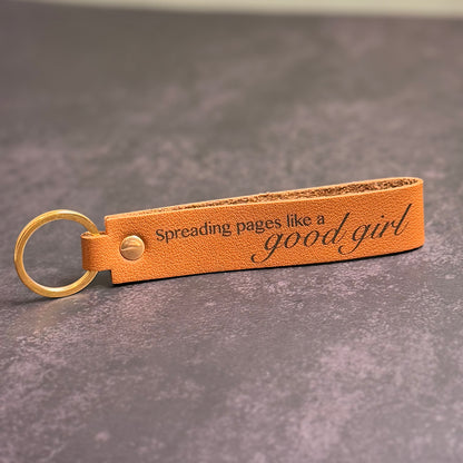A tan faux-leather keychain with 'Spreading pages like a good girl' etched into it.