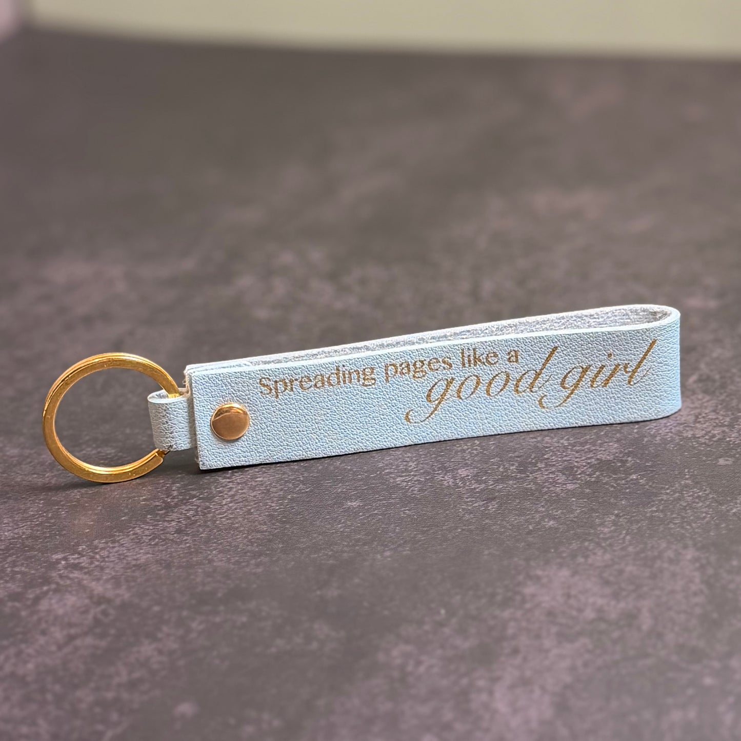 A sky blue faux-leather keychain with 'Spreading pages like a good girl' etched into it.