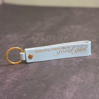 A sky blue faux-leather keychain with 'Spreading pages like a good girl' etched into it.