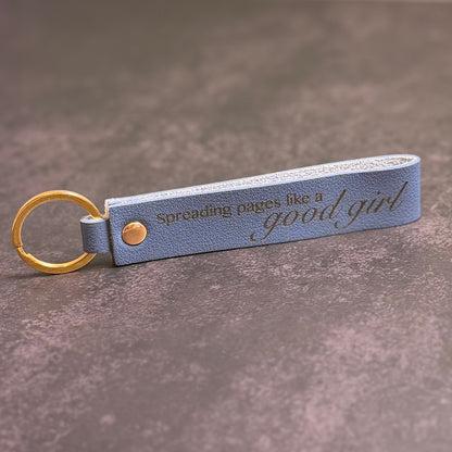 A navy blue faux-leather keychain with 'Spreading pages like a good girl' etched into it.