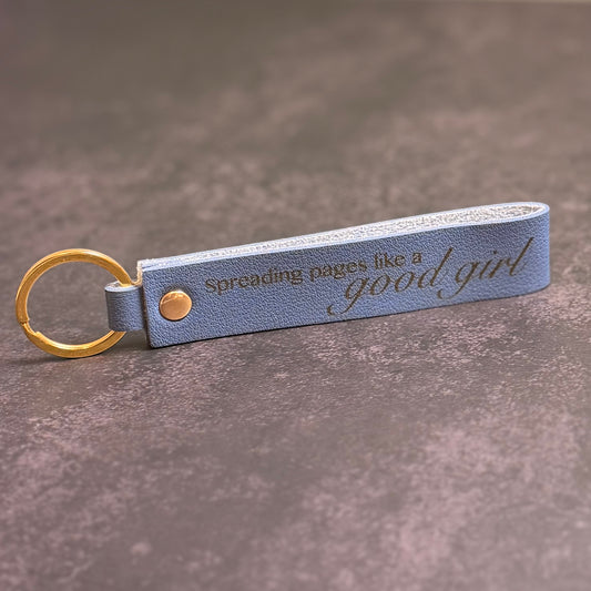 A navy blue faux-leather keychain with 'Spreading pages like a good girl' etched into it.