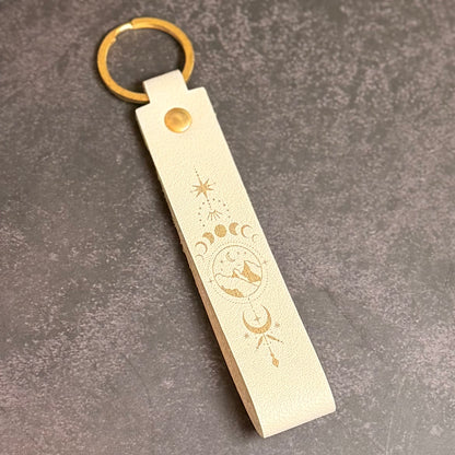 A eggshell white faux-leather keychain with moons and mountains etched into it.