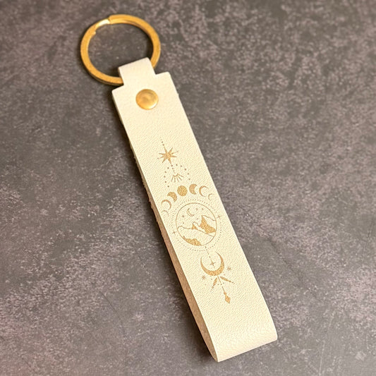 A eggshell white faux-leather keychain with moons and mountains etched into it.