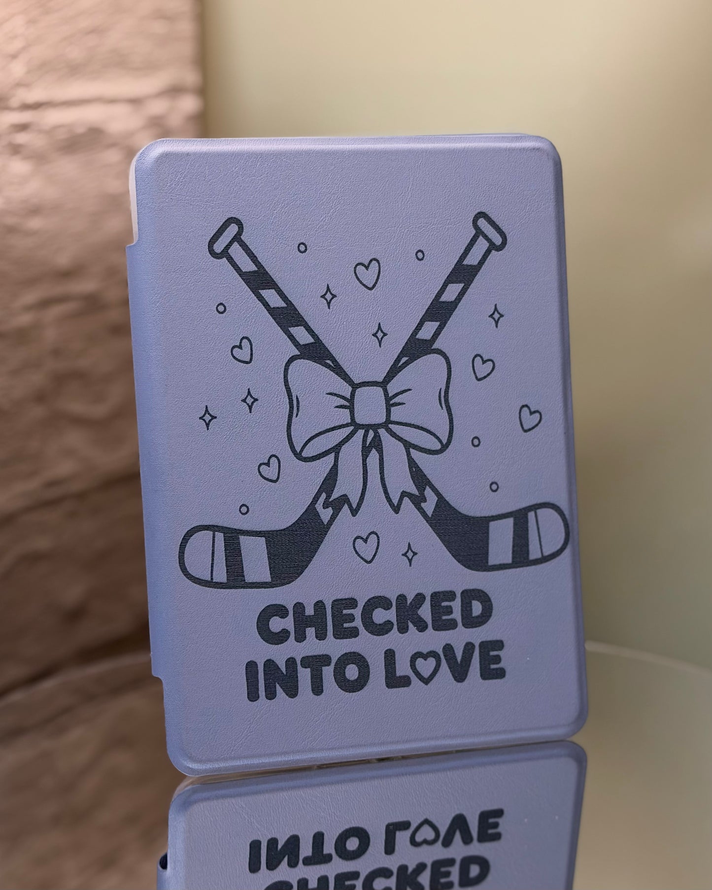 Dusty lavender Kindle case with hockey sticks and a bow, featuring the text 'Checked into Love' on a reflective surface. 