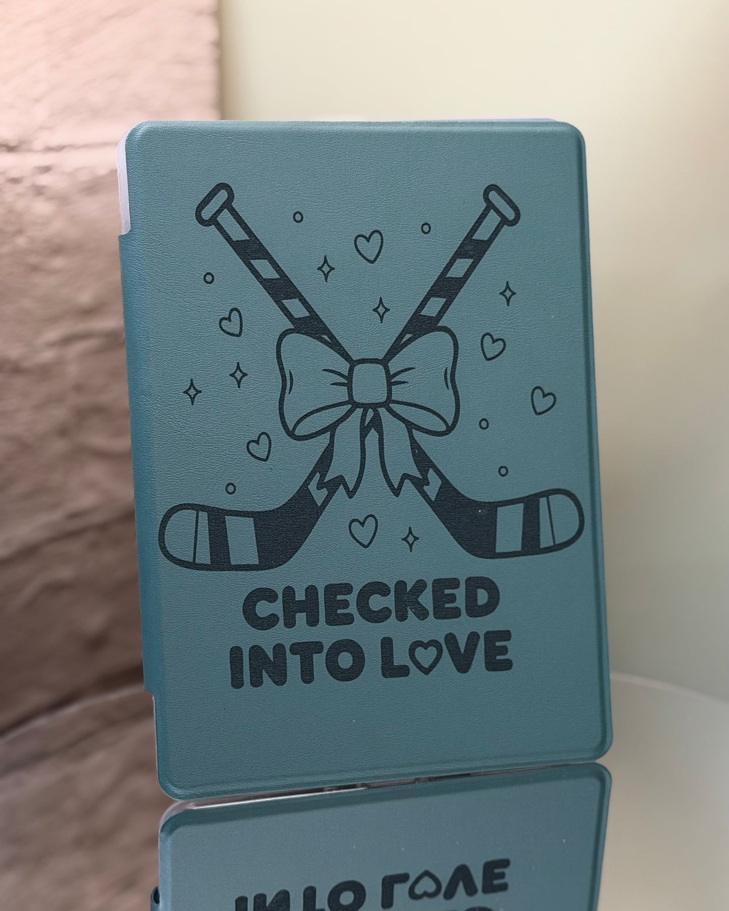 Dark sage Kindle case with hockey sticks and a bow, featuring the text 'Checked into Love' on a reflective surface. 