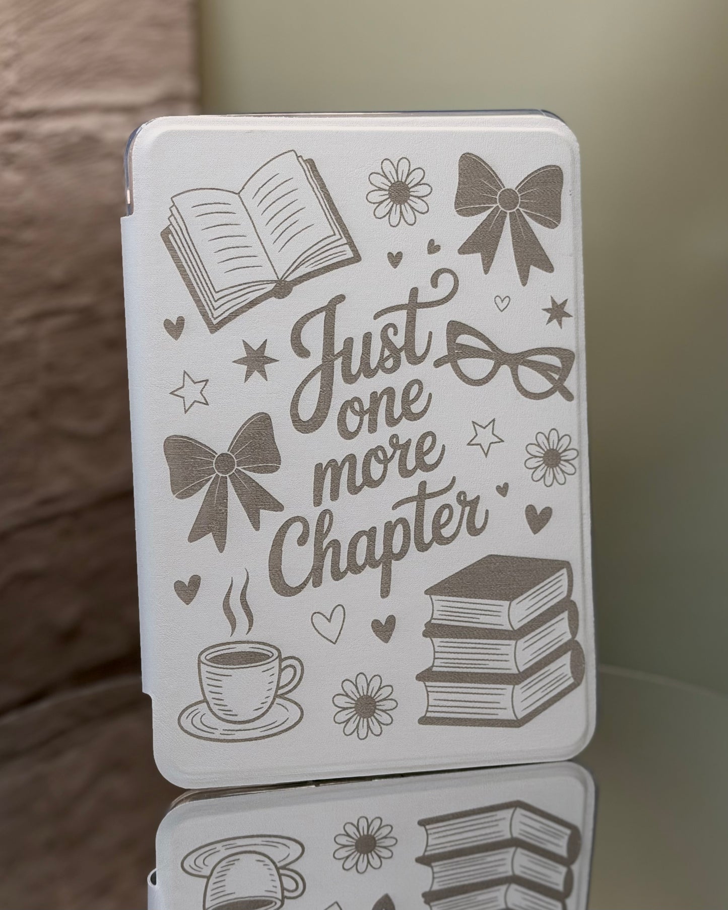 White Kindle case with engraved text saying just one more chapter and a variety of illustrations on a reflective surface