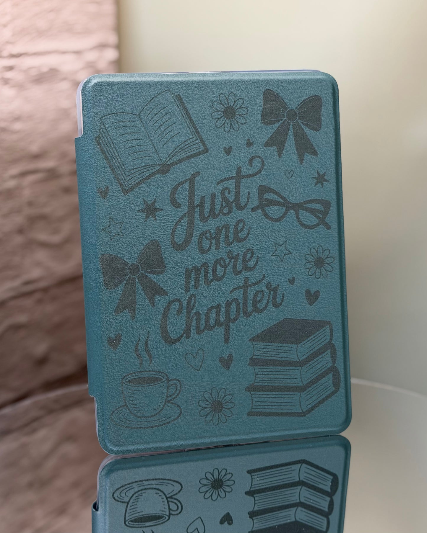 Dark sage Kindle case engraved with saying just one more chapter and a variety of illustrations on a reflective surface