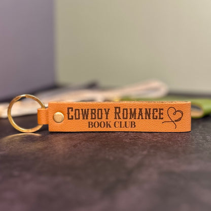 A brown faux-leather keychain with 'Cowboy romance book club' etched into it.