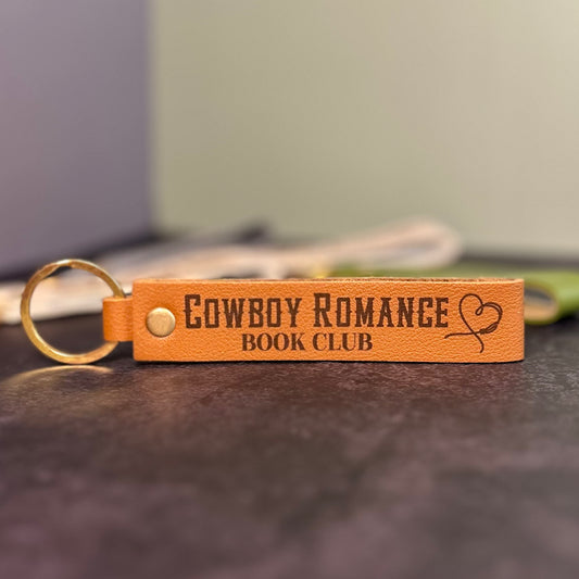 A brown faux-leather keychain with 'Cowboy romance book club' etched into it.