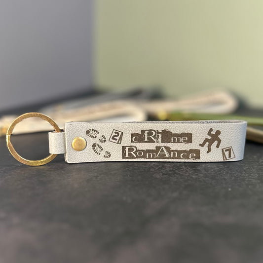 An eggshell white faux-leather keychain with 'Crime Romance' etched into it.